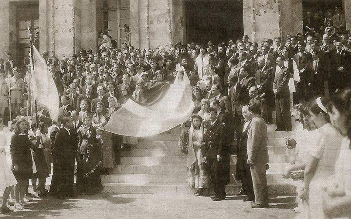 Ρόδος, 31 Μαρτίου 1947. Δώδεκα νέες με τοπικές ενδυμασίες που συμβολίζουν τα δώδεκα ελεύθερα νησιά σηκώνουν στα χέρια την ελληνική σημαία. Οι Αρχές και πλήθος κόσμου περιμένουν να ξεκινήσει η πομπή από το Δημαρχείο προς το κτήριο της Διοίκησης Δωδεκανήσου. Αρχείο Συμεών Δοντά