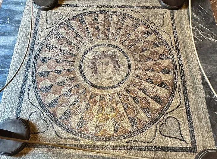 ΙΦΙΓΕΝΕΙΑ ΠΛΕΥΡΑΚΗ Ασπίδα με πολύχρωμη φολιδωτή διακόσμηση και κεντρικό έμβλημα με το πρόσωπο της Μέδουσας. Στις γωνίες μεταξύ του εμβλήματος και του ταινιωτού πλαισίου, φύλλα κισσού. Χρονολογείται γύρω στα τέλη του 3ου αιώνα με μέσα 4ου αιώνα.